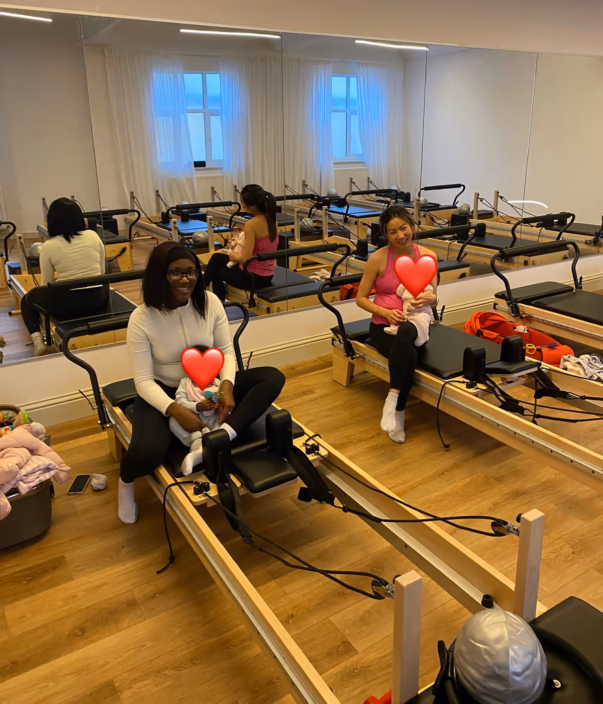 Two mums sitting on the reformers at Innerform's Lewisham studio, babies on their laps, during a Bump & Baby class.
