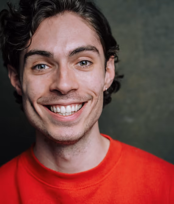 Jamie smiles broadly to camera in a red t-shirt, dark green backdrop.