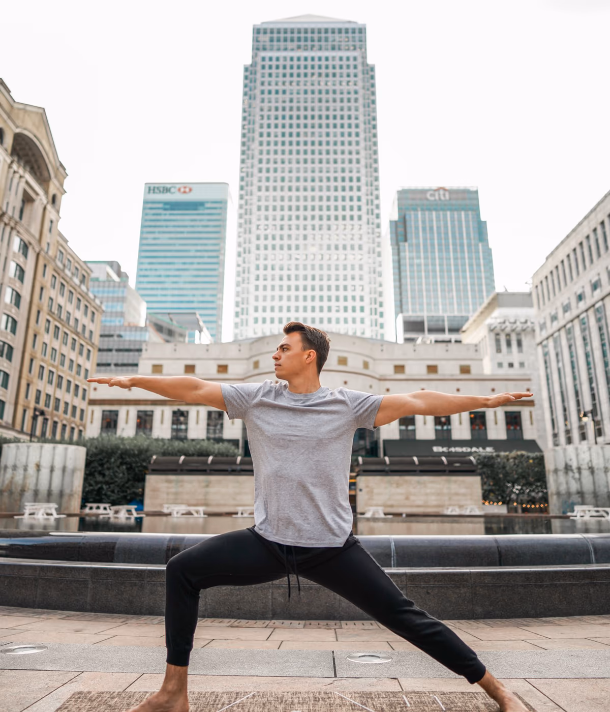 A teacher in Warrior II pose on a yoga mat outdoors, with the Canary Wharf skyline behind.